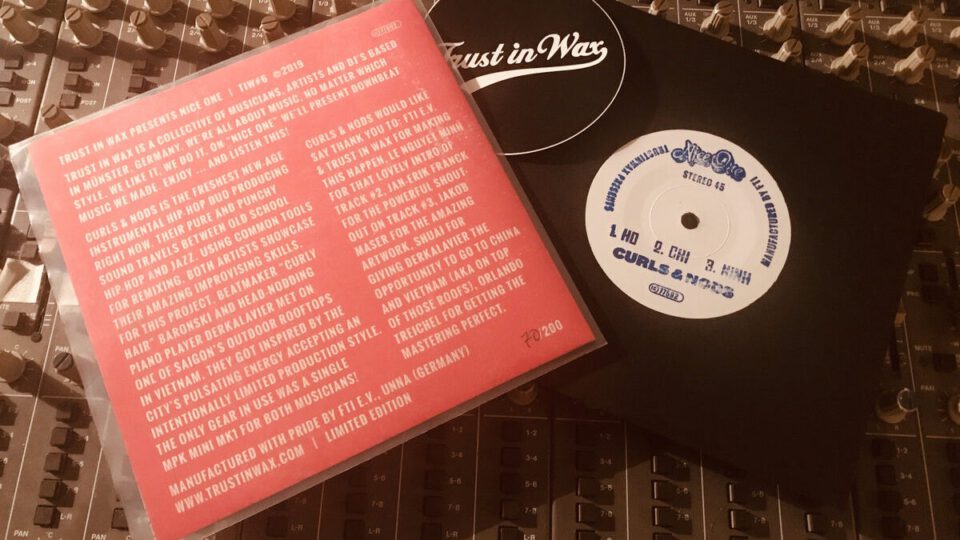 Schwarze Schallplatte mit weißem Label und rotem, englisch bedrucktem Cover auf einem Audiomischpult; daneben eine Hülle mit „Trust in Wax“.