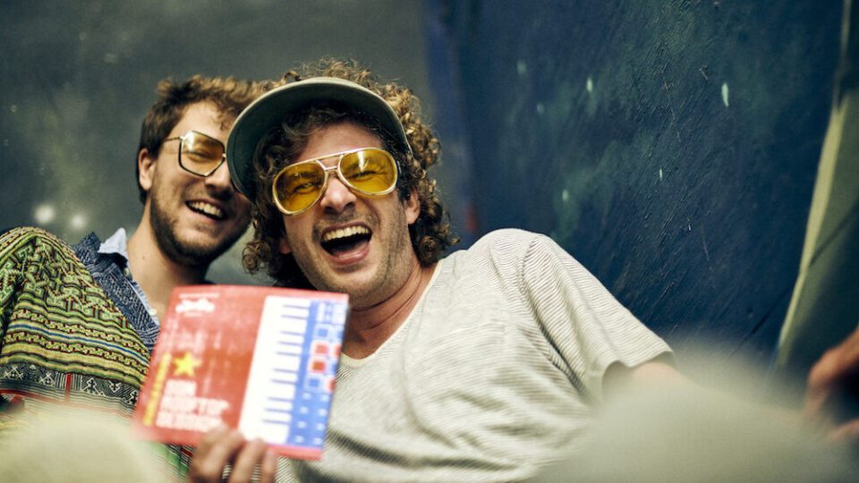 Zwei lachende Männer, einer mit Sonnenbrille und Hut, halten eine rote und blaue 7-Inch-Hülle in die Kamera.